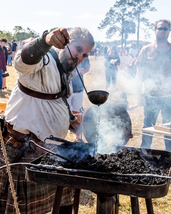 A blacksmith in a kilt works the forge as smoke rises and guests look on