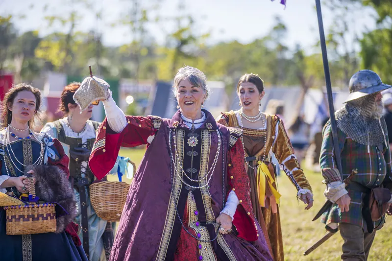 The Queen of Venezia in a purple and gold gown waves as she leads her ladies-in-waiting and a helmeted guard through the faire