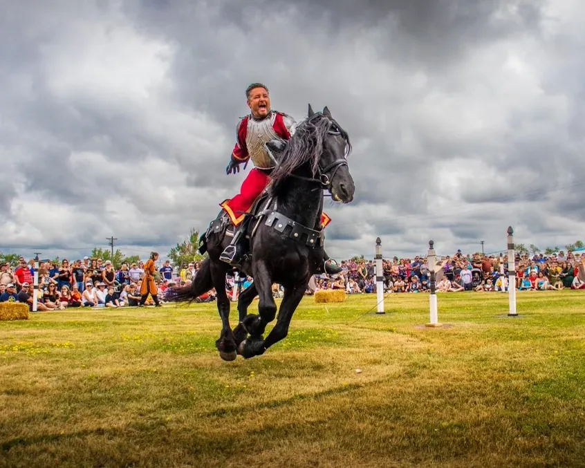 A Dark Knight in red and silver armour gallops a black horse past the cheering tournament crowd
