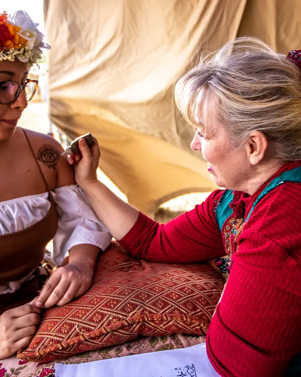 A girl with a flower crown has her face painted by an artist in a red bodice