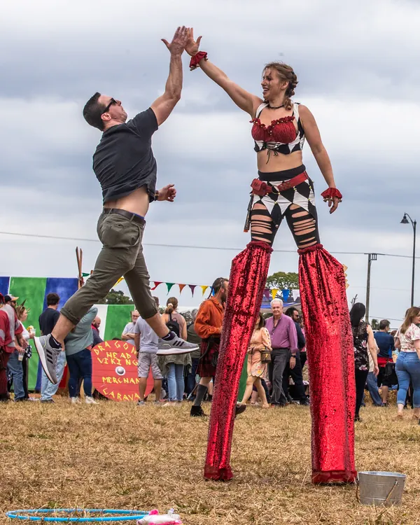 A stilt-walker in red high-fives a guest in the middle of the faire field